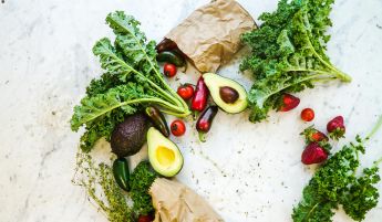 Avocado, kale, and more fresh produce artfully arranged on a marble surface.